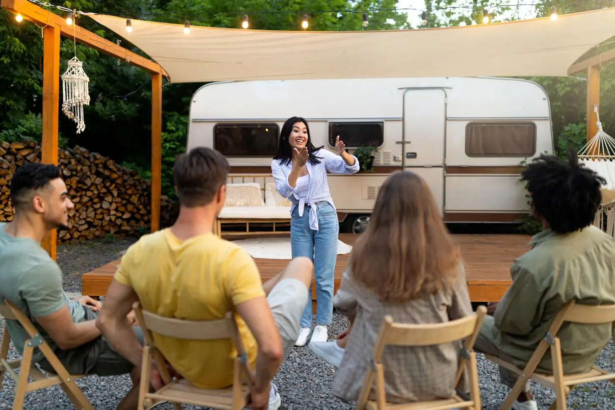 A woman plays charades with her friends outside their RV under a canopy with string lights.