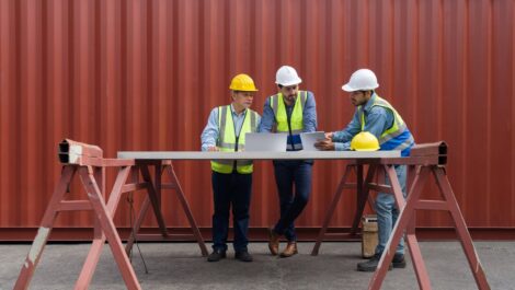 Three construction workers in safety gear review blueprints at a table in front of a large red storage container.